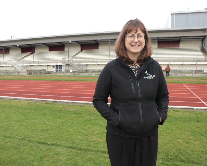 A woman stands in an athletics field in front of a grandstand. 