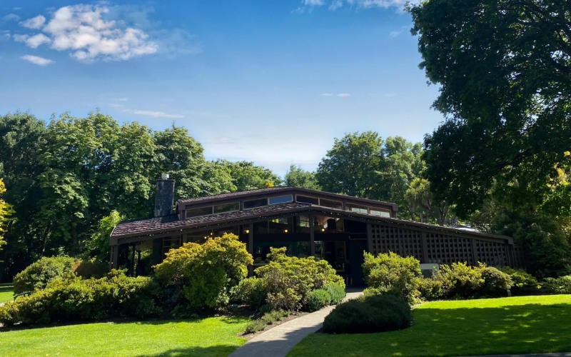 A low mid-century style building sits among mature trees, garden plants, and green lawn.