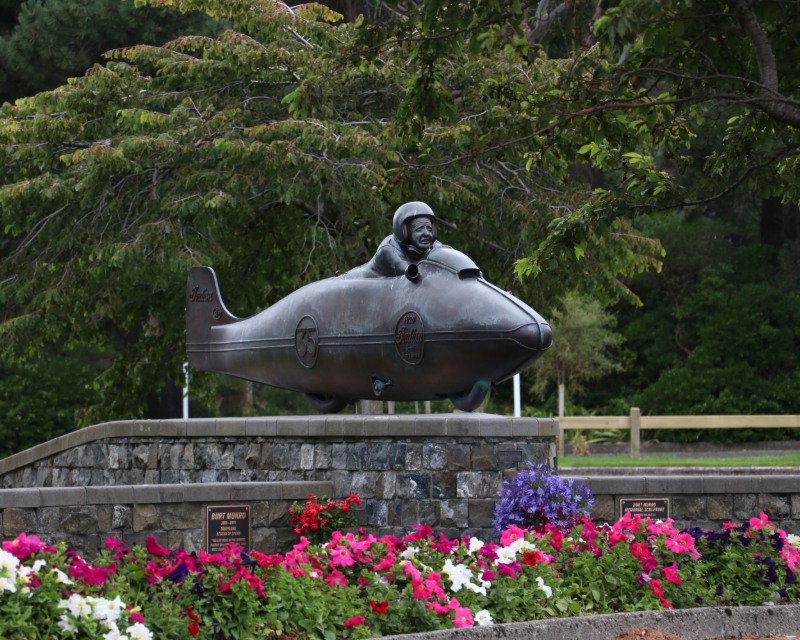 A sculpture of Burt Munro in a vehicle sits on a stone foundation in front of a flowering garden with trees in the background.