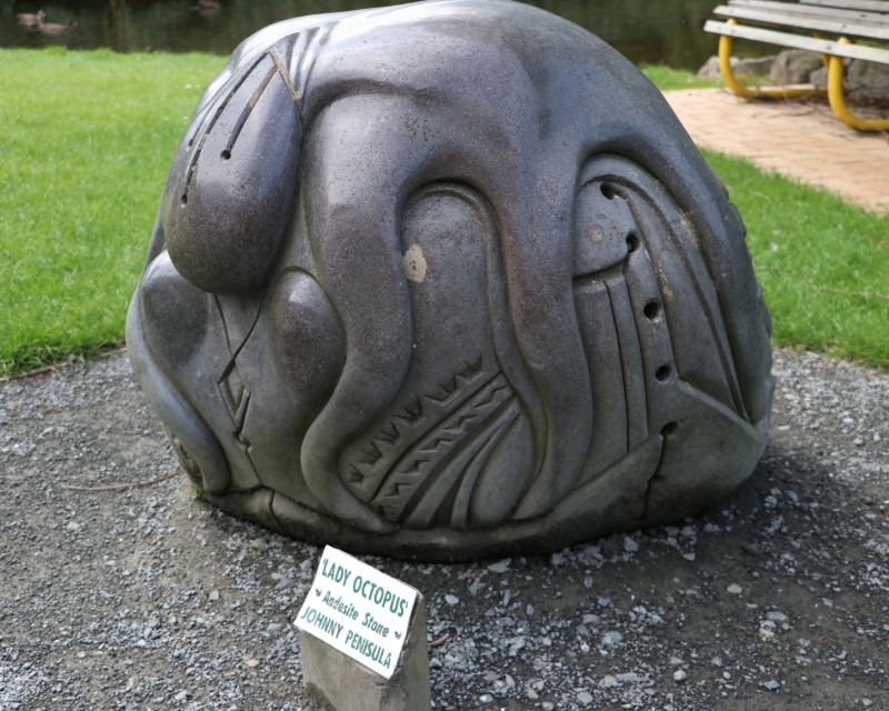 A grey stone sculpture in a round boulder shape with a carved octopus-like figure draped over it. 