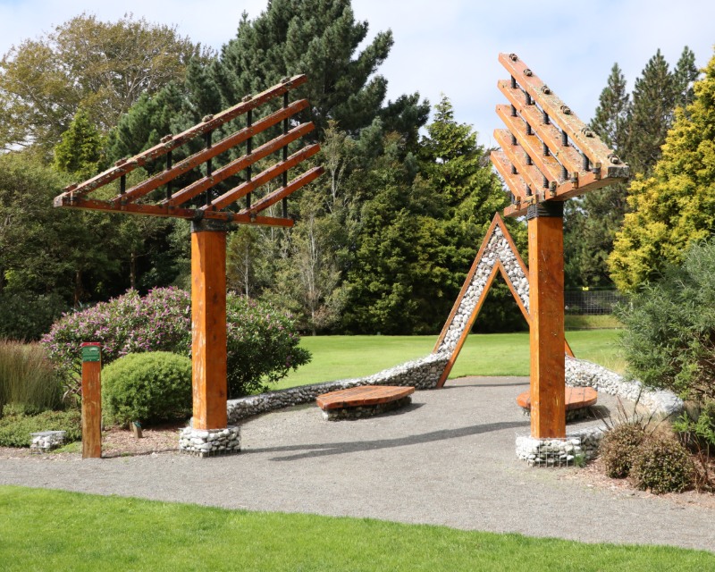 A wooden sculpture made up of triangle shapes sits on a paved path among the lawns and trees of Queens Park.