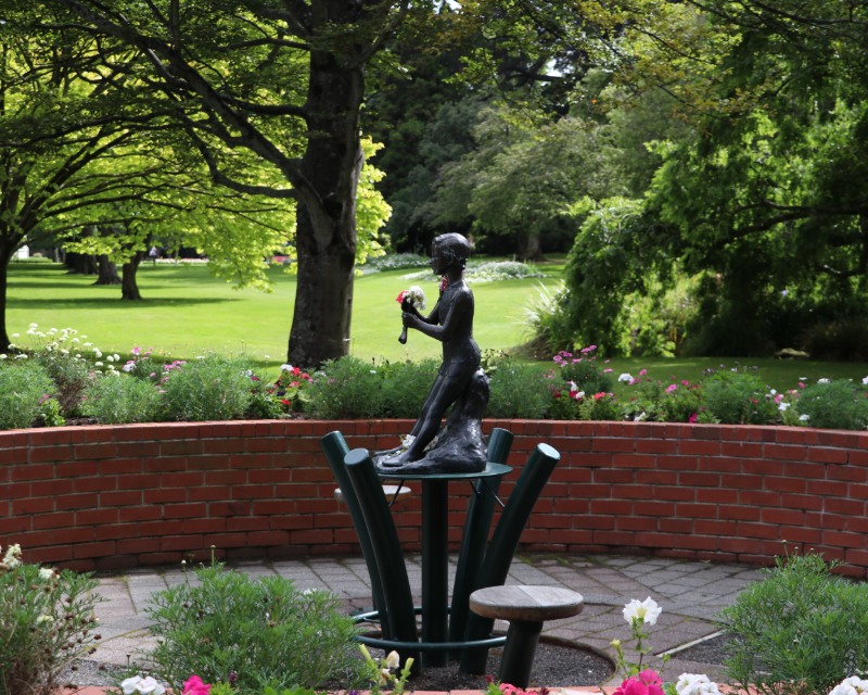 A statue of Peter Pan sits on a paved area among planted gardens, with a low brick wall, trees, and lawn behind.