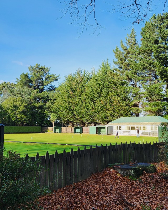 A small white pavilion surrounded by trees fronts onto a green croquet lawn bounded by a picket fence. 