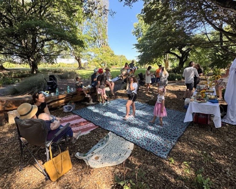 Children playing under trees on a picnic blanket among logs and woodchips.