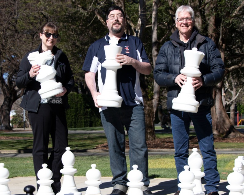 Three people stand side by side holding large white chess pieces in a park.