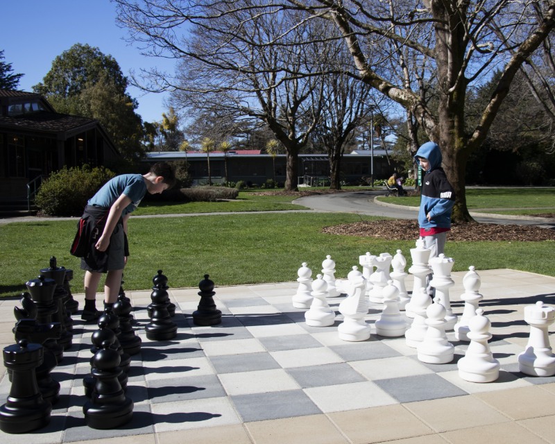 Two boys play chess on a giant chess board in a park.