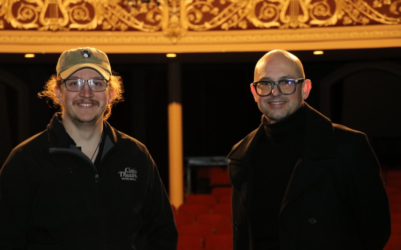 Two men stand in front of the ornate decorations inside a theatre.