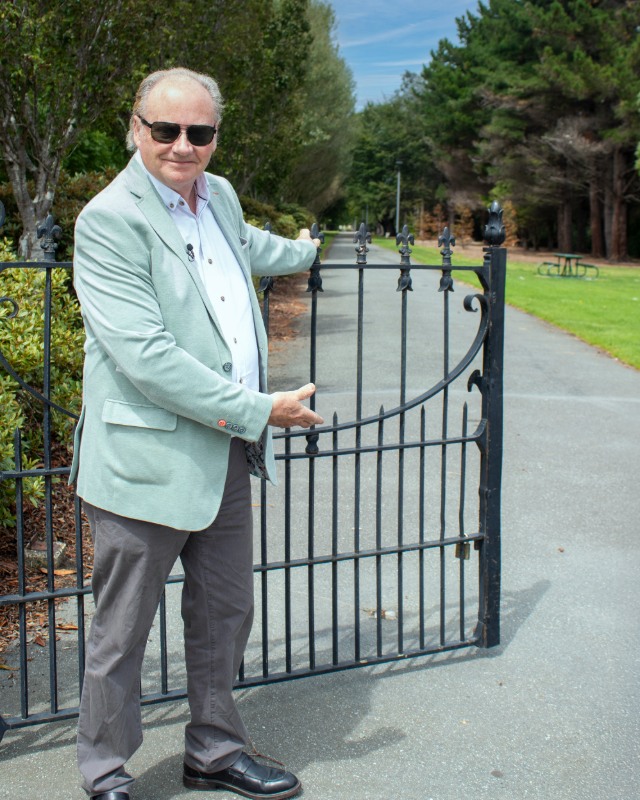 A man stands opening a gate leading to a path into a park with grass and trees.
