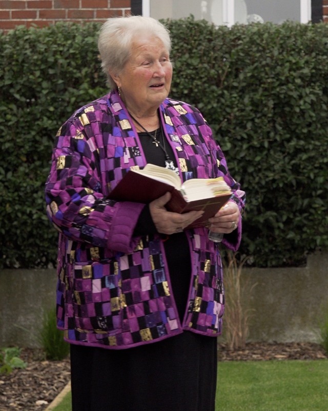A woman with a purple and punk patterned coat holds a large book while looking in front of her. There is a green leafy hedge in the background and grass is on the ground. 