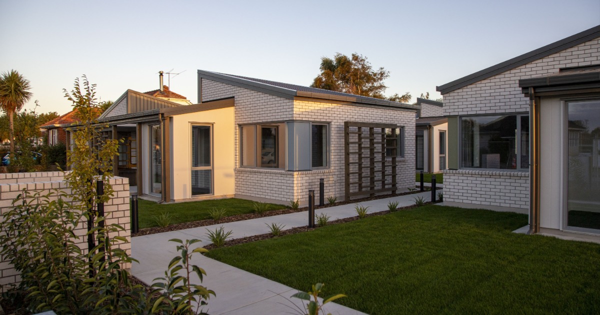 Exterior view of a newly completed single-level housing complex with several modern brick units arranged around a shared lawn and paved walkways, shown in warm late-afternoon light.