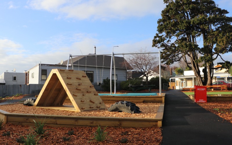 Wooden climbing structure in a landscaped playground, featuring an A-frame climbing wall with handholds and a woodchip safety surface. The play area is bordered by timber edging and connected by accessible pathways. Trees, nearby buildings and park infrastructure are visible in the background, highlighting a well-designed community recreation space that encourages active outdoor play. 