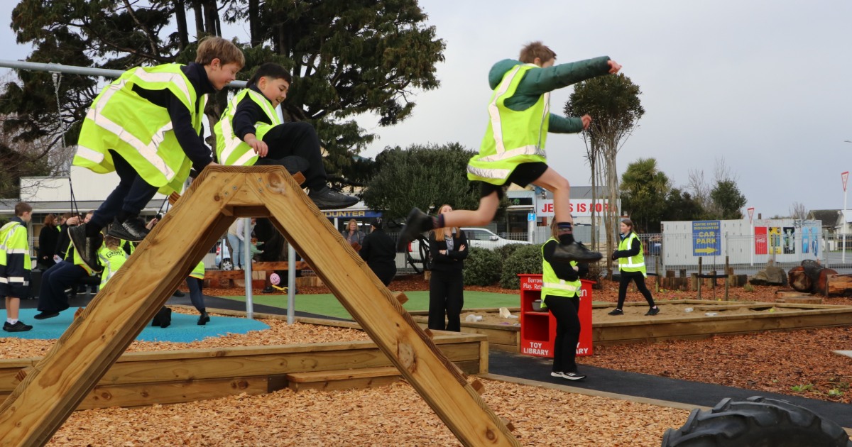 Children wearing high-visibility vests enjoy an outdoor adventure playground, climbing and jumping across timber obstacle structures. One child is captured mid-air between wooden frames while others balance and climb nearby. The woodchip-covered play area is designed to encourage active play, coordination and confidence in a safe outdoor environment. Trees, pathways and additional play elements are visible in the background.