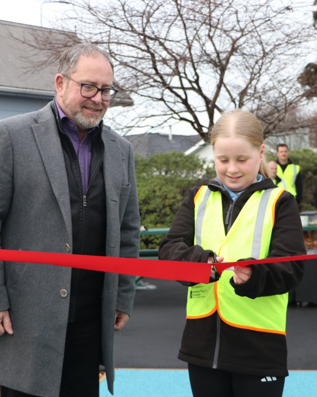 A person wearing a high-visibility safety vest cuts a red ceremonial ribbon with scissors during an outdoor opening event. Another person stands beside the ribbon-cutting participant near a paved area and playground. Trees, buildings and several people are visible in the background, highlighting a community celebration marking the opening of a new public space or facility.
