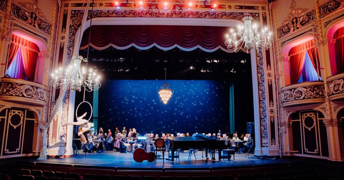 Interior of the Civic Theatre showing guests seated at tables on stage beneath chandeliers, with a grand piano, double bass and aerial performer in front of a blue, star-lit backdrop, framed by the theatre’s ornate historic architecture.